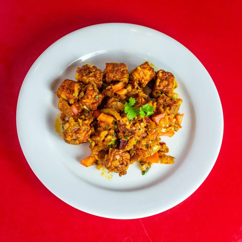 A white plate of Indian chili paneer, garnished with cilantro, on a red background.