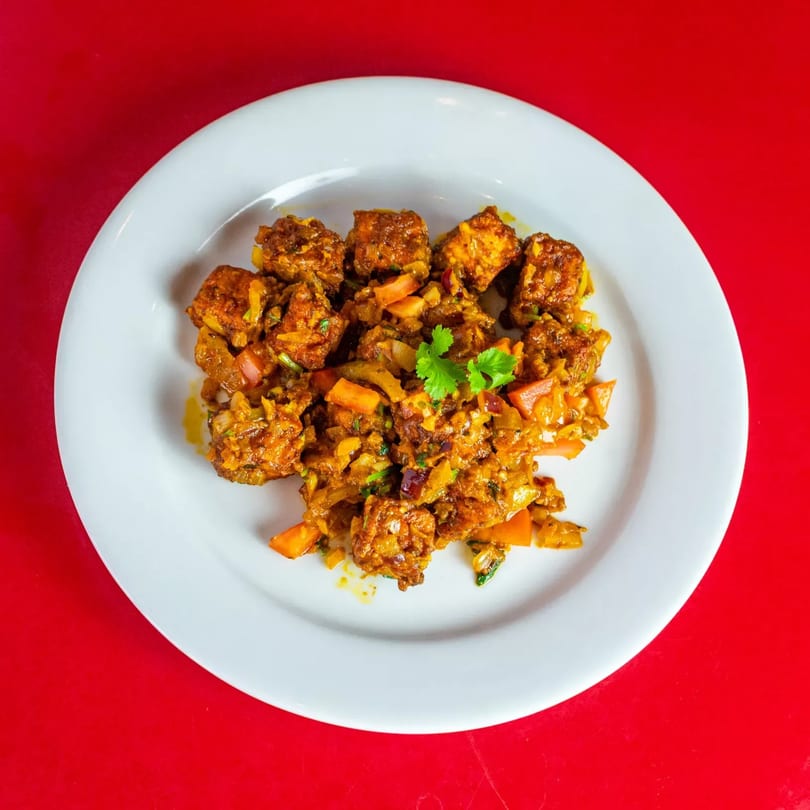 A white plate of Indian chili paneer, garnished with cilantro, on a red background.